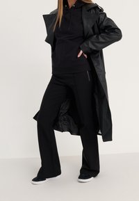 Gabardina de cuero negro sobre una sudadera con capucha negra y pantalones negros. El conjunto presenta un diseño minimalista y un ajuste relajado, combinado con zapatillas oscuras.