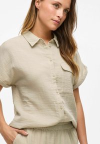 Camicia beige chiaro a maniche corte con bottoni realizzata in tessuto testurizzato, con taschino sul petto e un design a vestibilità rilassata.