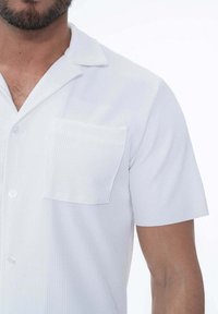 Chemise légère à manches courtes en blanc avec un motif rayé vertical subtil. Équipée d'un col en V et d'une poche plaquée sur la poitrine gauche.