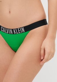 Žalios thong kelnaitės su juodu elastiniu juosmeniu, ant kurio užrašyta "CALVIN KLEIN" baltomis raidėmis. Lygus paviršius ir minimalistinis dizainas.