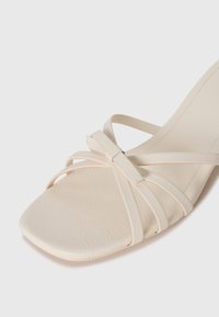 Beige stroppesandaler med firkantet tå, teksturert lærøvre og en knutedetalj i midten. Glatt, stilren hældesign.