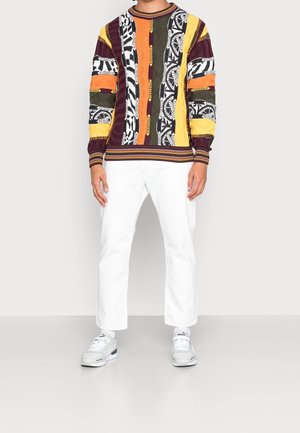 Meer kleurige gebreide pullover met gestreepte patronen in oranje, geel, zwart en wit, gecombineerd met witte broek en grijze sneakers.