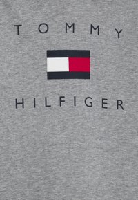 Grauer Pullover mit dem Logo "TOMMY HILFIGER" in fettgedruckten schwarzen Buchstaben, mit einer rechteckigen, mehrfarbigen Flagge Grafik darüber.