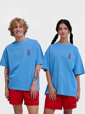Młody mężczyzna i kobieta noszący pasujące do siebie niebieskie, oversize'owe koszulki i czerwone szorty, stojący obok siebie na jednolitym tle.
