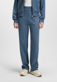 Blue athletic pants in a smooth fabric feature a tapered design with side seams and a comfortable waistband, paired with beige sneakers.