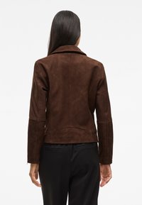 Brun mockasinsjakke med en tettsittende design, skjært krage og lange ermer. Har paneldetaljer og en litt forkortet lengde.