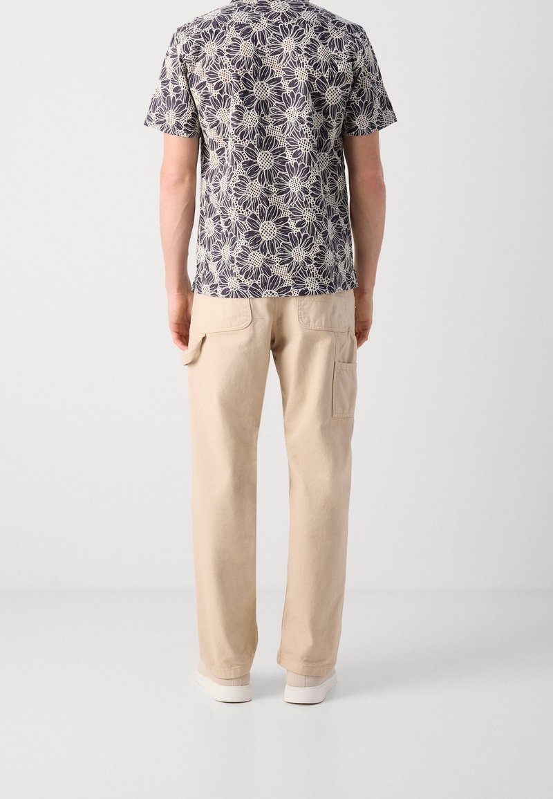 Homme portant un pantalon cargo beige, des baskets blanches et une chemise à manches courtes noire et blanche à motif floral, debout devant un fond clair uni.