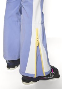 Paarse en witte skibroek met een zijrits en gele accenten, gemaakt van een glad, flexibel materiaal. Zwarte skibotten zijn gedeeltelijk zichtbaar.