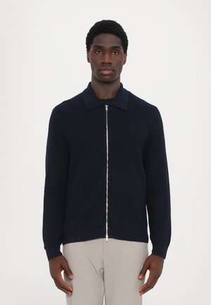 Pulover bleumarin cu nervuri, cu fermoar în față, având un guler ascuțit și mâneci lungi. Fabricat dintr-un material texturat, are un design ajustat.