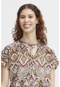 Un chemisier à manches courtes avec un motif paisley multicolore dans des tons de marron, vert et lavande, avec un détail à nouer au niveau du col.