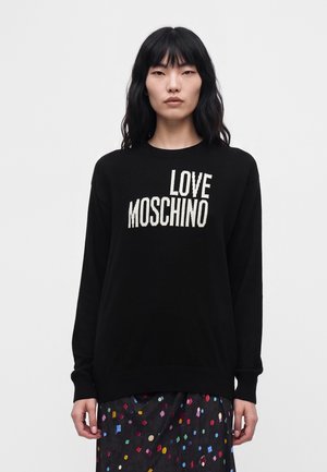 Жена с черна коса, облечена в черен пуловер с текст "LOVE MOSCHINO" и черна пола с шарки, приличащи на цветни конфети.