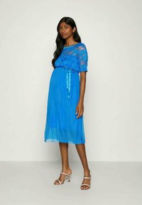 Plava čipkasta haljina za trudnice s kratkim rukavima, uskim korzetom i lepršavom suknjom do koljena. Završena s satenskom mašnom u struku.