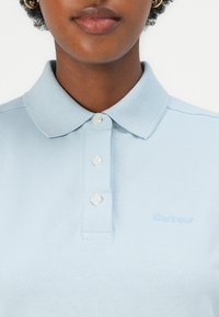 Hele sinine puuvillane polo-särk tekstuurse kangaga, kolme nupuga krael ja broderitud "Barbour" logo sama tooni.