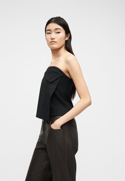 Γυναίκα με μακριά σκούρα μαλλιά, φορώντας μαύρο τοπ χωρίς τιράντες και σκούρο, ψηλόμεσο παντελόνι, με το ένα χέρι στην τσέπη, κοιτάζοντας πλάγια μπροστά σε λευκό φόντο.