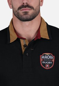 Polo shirt de rugby noir avec un col marron et rouge, doté d'un logo brodé, de trois boutons et de détails de couture contrastants.