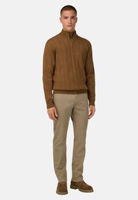 Brązowy sweter dzianinowy z wysokim kołnierzem i zamkiem, zdobiony wzorem liści. W połączeniu z jasnobeżowymi spodniami i brązowymi zamszowymi butami.