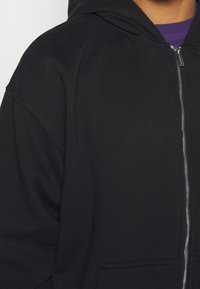 En person som bär en svart zip-up hoodie över en lila tröja, dragkedjan delvis stängd, synlig från axeln till mitten av bröstet.