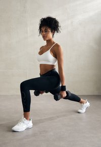 Leggings negros con el texto "NIKE PRO", un sujetador deportivo blanco y zapatillas deportivas blancas; sosteniendo dos mancuernas negras, en posición de zancada.
