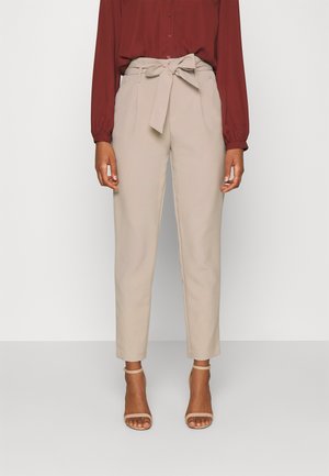 Beżowe spodnie na miarę z paskiem wiązanym w talii, o dopasowanym kroju i gładkiej fakturze. Połączone z bordową koszulą na guziki i sandałkami na paski.