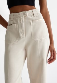 Beige, højtaljede bukser i et tekstureret stof med to baglommer og metalliske detaljer. Figursyet pasform med en glat overflade.