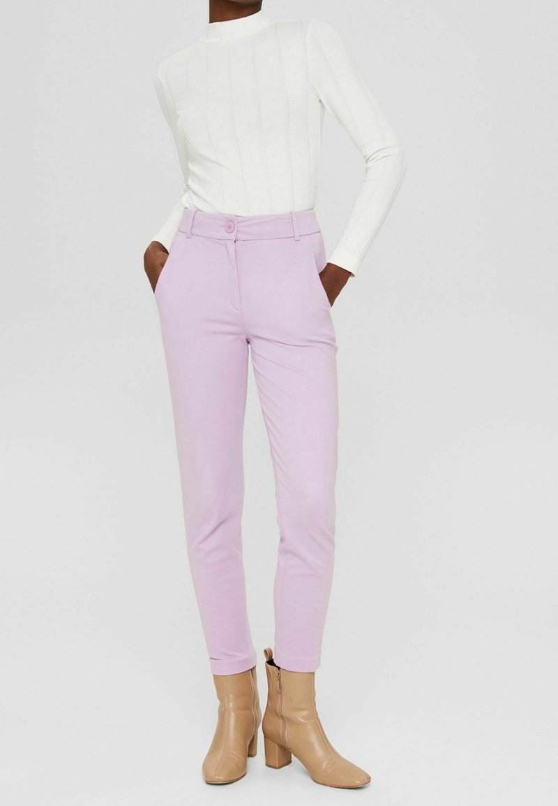 Personne portant un pantalon ajusté violet pâle, un haut blanc à manches longues et des bottines beige, debout avec les mains dans les poches.