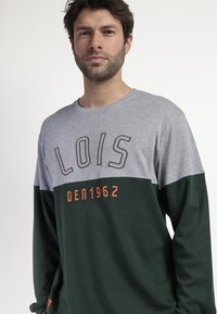 Camiseta de manga larga, la parte superior gris y la parte inferior verde. Presenta bordados en color naranja con el texto "LOIS" y "DEN1962". Material de algodón suave.