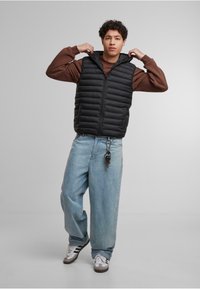 Čierna prešívaná vesta s horizontálnym prešívaním, so zipsom a kapucňou, zkombinovaná s hnedým sveterom s okrúhlym výstrihom a svetlomodrými voľnými džínsami.