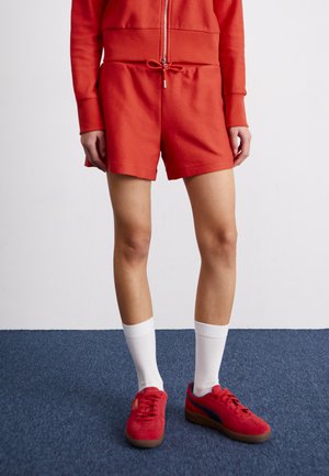 Pantalones cortos deportivos rojos con cintura ajustable, combinados con zapatillas rojas y calcetines blancos hasta el tobillo. La tela tiene un patrón texturizado.