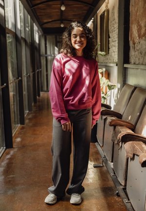 Roze sweatshirt, donkergrijze broek en witte sneakers. De outfit heeft een ontspannen pasvorm en is gemaakt van zachte, textuurvolle materialen.