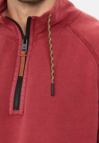 Sweat-shirt bordeaux avec fermeture à moitié éclair, doté d'une fermeture éclair noire, d'une languette en cuir et d'un cordon de serrage à motif orange et noir.