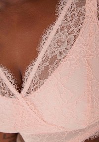 Nahaufnahme eines Oberkörpers mit blassrosa floralem Spitzen-BH mit gewellten Rändern und filigranen Netz-Details.