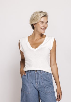 Smiling woman with short blonde hair wearing a white sleeveless top and high-waisted blue denim pants with hands in pockets.