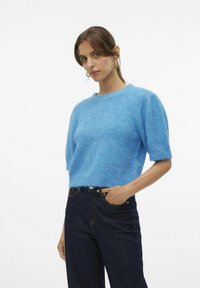Maglione blu peloso con maniche corte, scollo rotondo e vestibilità comoda, abbinato a jeans in denim scuro. Materiale morbido e testurizzato.