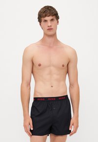 Boxer-short noirs avec une ceinture rouge portant la marque "HUGO", dotés d'une fermeture à bouton et d'une texture de tissu lisse et légère.