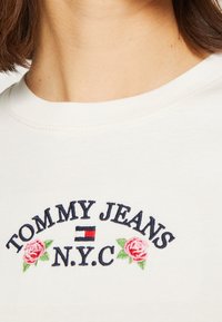 Bílá bavlněná tričko s vyšitým textem "TOMMY JEANS N.Y.C" v námořnické modré a červené barvě, s růžovými akcenty růží po stranách.