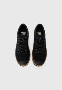 Ein Paar schwarze Wildleder-Wanderschuhe mit schwarz-weißen Schnürsenkeln, braunen Gummisohlen und bedruckten Einlegesohlen vor einem weißen Hintergrund.