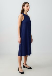 Vrouw die zijwaarts staat in een mouwloze marineblauwe midi-jurk en zwarte lage Mary Jane-schoenen tegen een eenvoudige lichte achtergrond.