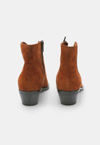Botas de tobillo de ante marrón con un tacón negro bajo, que cuentan con una cremallera lateral y una textura suave. El diseño es elegante y ajustado.
