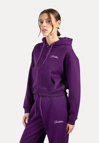 Lilla zip-up hættejakke med frontlomme, elastiske manchetter og matchende svedbukser. Har broderet logo på brystet og en blød stofstruktur.