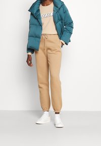 Turkusowa kurtka puchowa, beżowy T-shirt z grafiką oraz beżowe dresy z elastycznymi mankietami. Białe sneakersy dopełniają strój. Styl luźnej wygody.