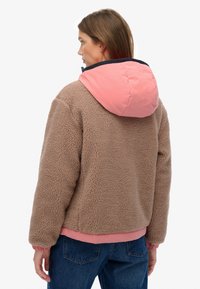 Tan faux fur jacket with a pink nylon hood, elastic cuffs, and hem. The design features a textured surface and a contrasting navy trim.