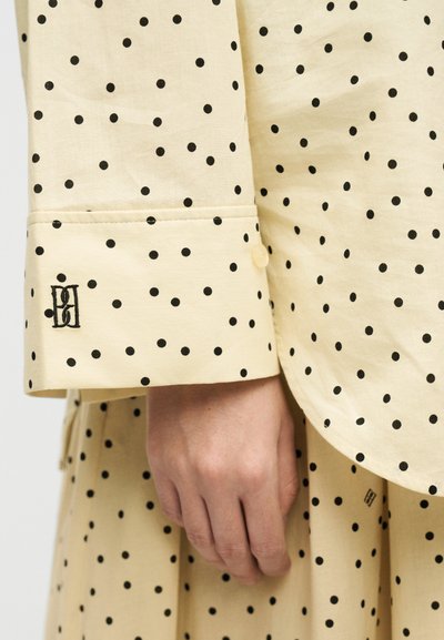 Χέρι που ακουμπά δίπλα σε μπεζ ύφασμα με μαύρες πουά, με κεντημένο μαύρο μονόγραμμα και μανσέτα με κουμπί.