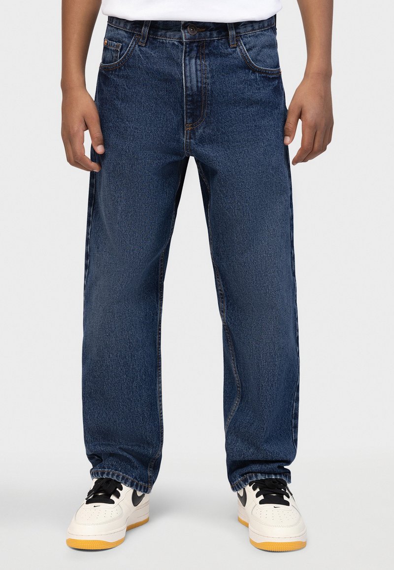 A kék farmernadrág egyenes szárú kialakítású, ötpántos stílusú, finoman kifakult, fehér tornacipőkkel és fekete részletekkel párosítva.