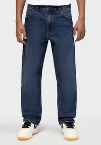 Τα μπλε τζιν διαθέτουν σχέδιο με ίσια γραμμή, στιλ με πέντε τσέπες και διακριτική ξεθώριαση, συνδυασμένα με λευκά αθλητικά παπούτσια και μαύρες λεπτομέρειες.