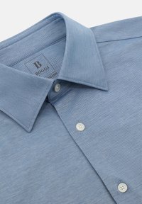 Camicetta azzurro chiaro con bottoni realizzata in un tessuto texturizzato, caratterizzata da un colletto classico, due bottoni bianchi sul davanti e un'etichetta con logo.