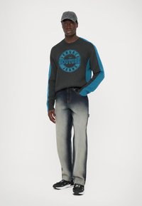 Grauer Pullover mit blauen Akzenten und Logo, kombiniert mit ausgewaschenen blauen Jeans. Das Modell trägt eine Mütze und schwarze Sneakers. Einfaches Design und entspannter Schnitt.