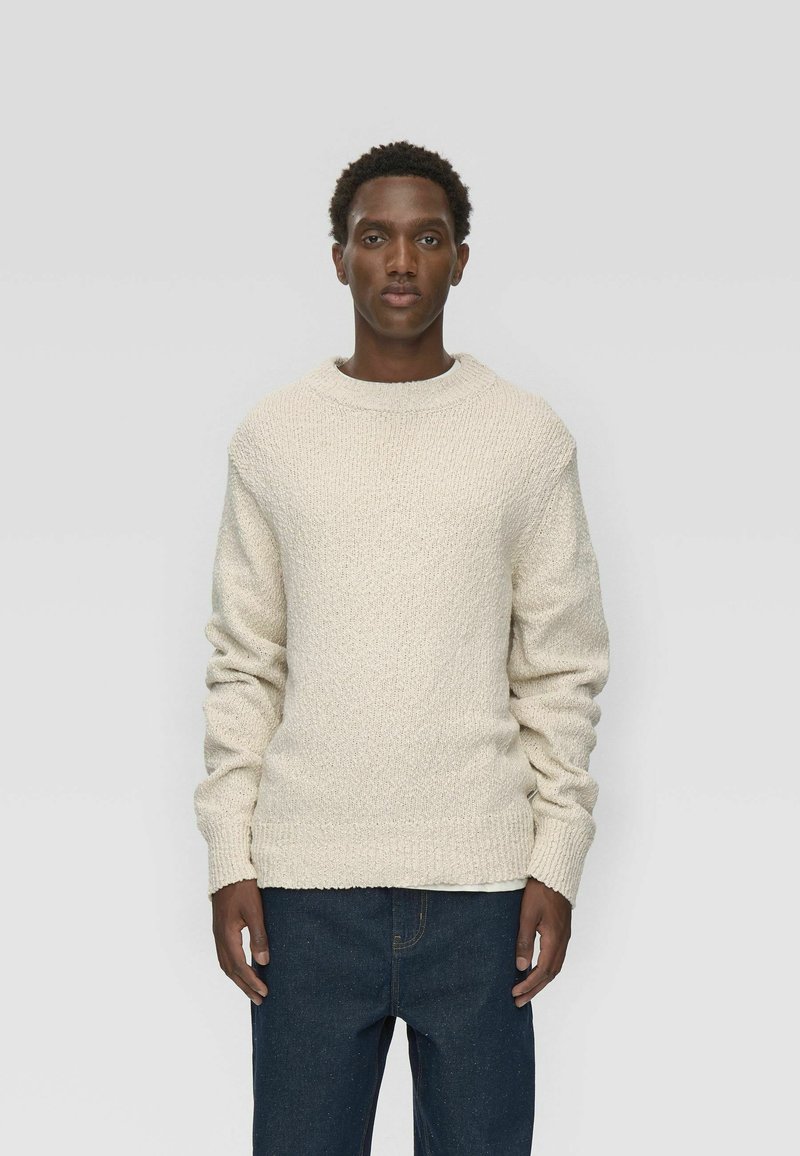 Pull en tricot crème à motif texturé, col rond et poignets côtelés, associé à un jean bleu foncé.