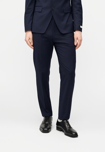 Σκούρα μπλε παντελόνια φόρμας από απαλό ύφασμα, με εφαρμοστή ραφή και διακριτική πτυχή, σε συνδυασμό με στιλβωμένα μαύρα δερμάτινα παπούτσια.