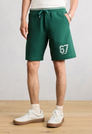 Uomo che indossa pantaloncini verdi con coulisse con la scritta "67 Marc O'Polo", calzini bianchi e scarpe da ginnastica bianche, in piedi su pavimento in legno.