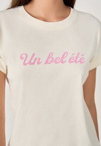 Camiseta de rayas crema con mangas cortas que presenta un texto rosa que dice "Un bel été". Textura suave y escote redondo.